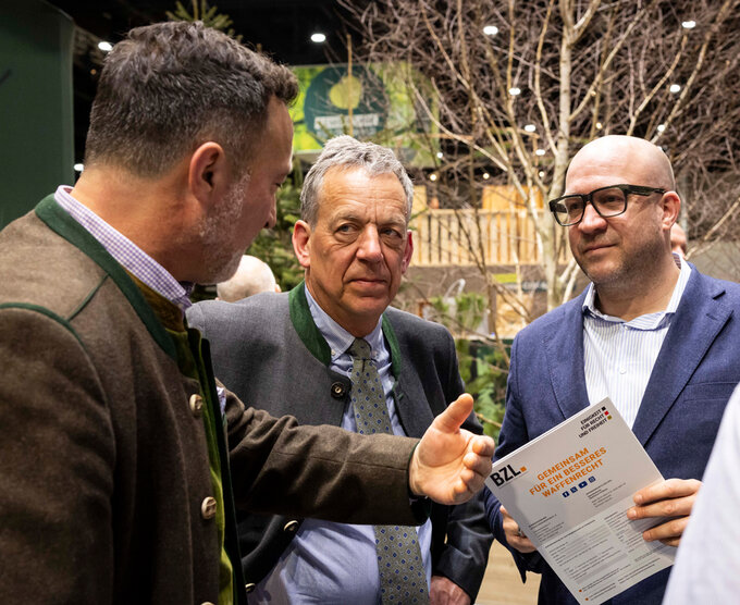 Drei Männer stehen im Gespräch bei einer Veranstaltung. Einer hält ein Informationsblatt in der Hand, während ein anderer gestikulierend etwas erklärt. Im Hintergrund sind Pflanzen und ein Messeumfeld zu sehen.