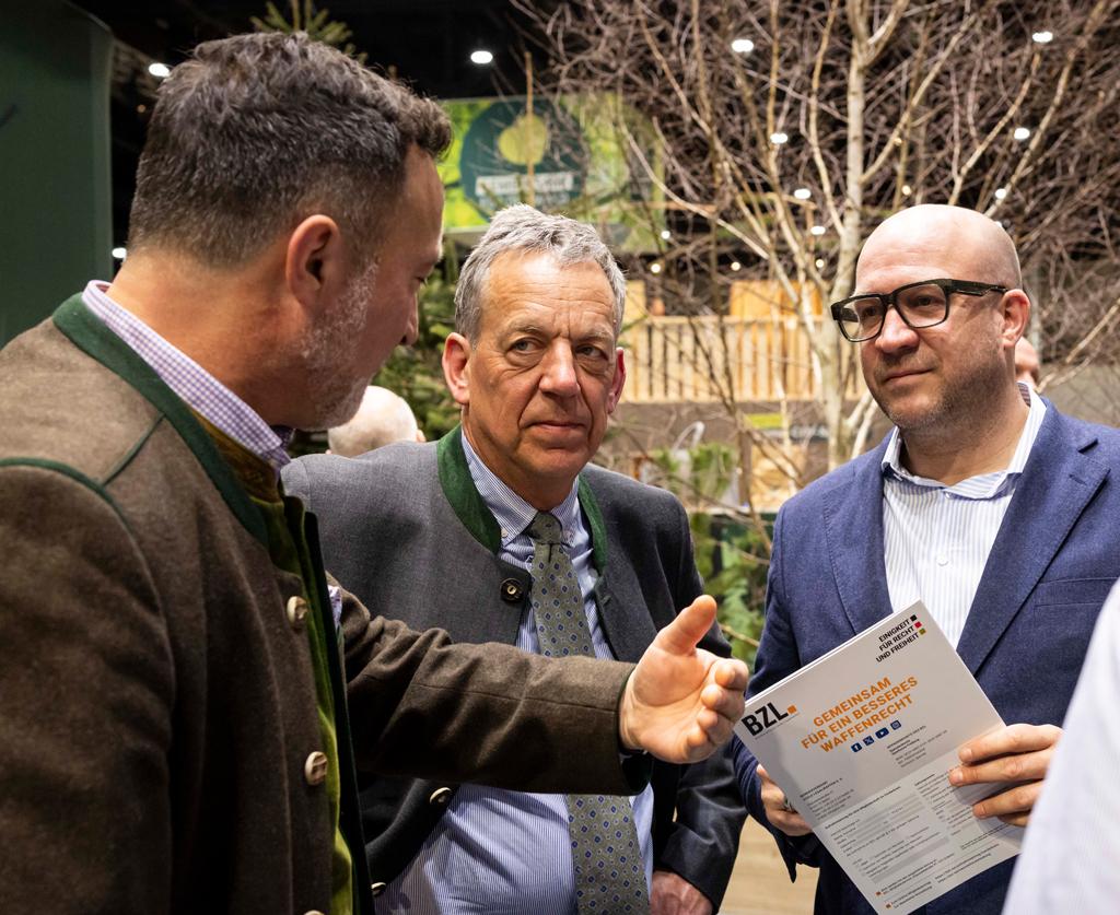 Drei Männer stehen im Gespräch bei einer Veranstaltung. Einer hält ein Informationsblatt in der Hand, während ein anderer gestikulierend etwas erklärt. Im Hintergrund sind Pflanzen und ein Messeumfeld zu sehen.